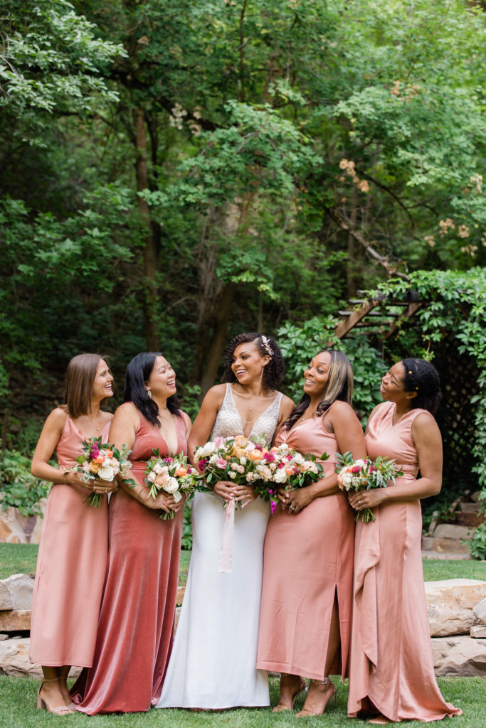 Utah bridesmaids party smiling and posing for a photo surrounded by rich greens and colorful flowers. Lush Mountain Wedding