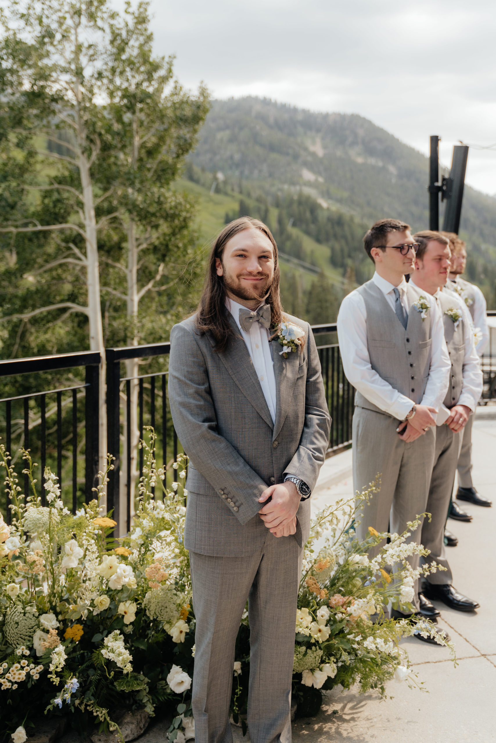 A groom stands in front of a nest of wildflowers smiling while watching his bride walk down the aisle with a mountain backdrop.