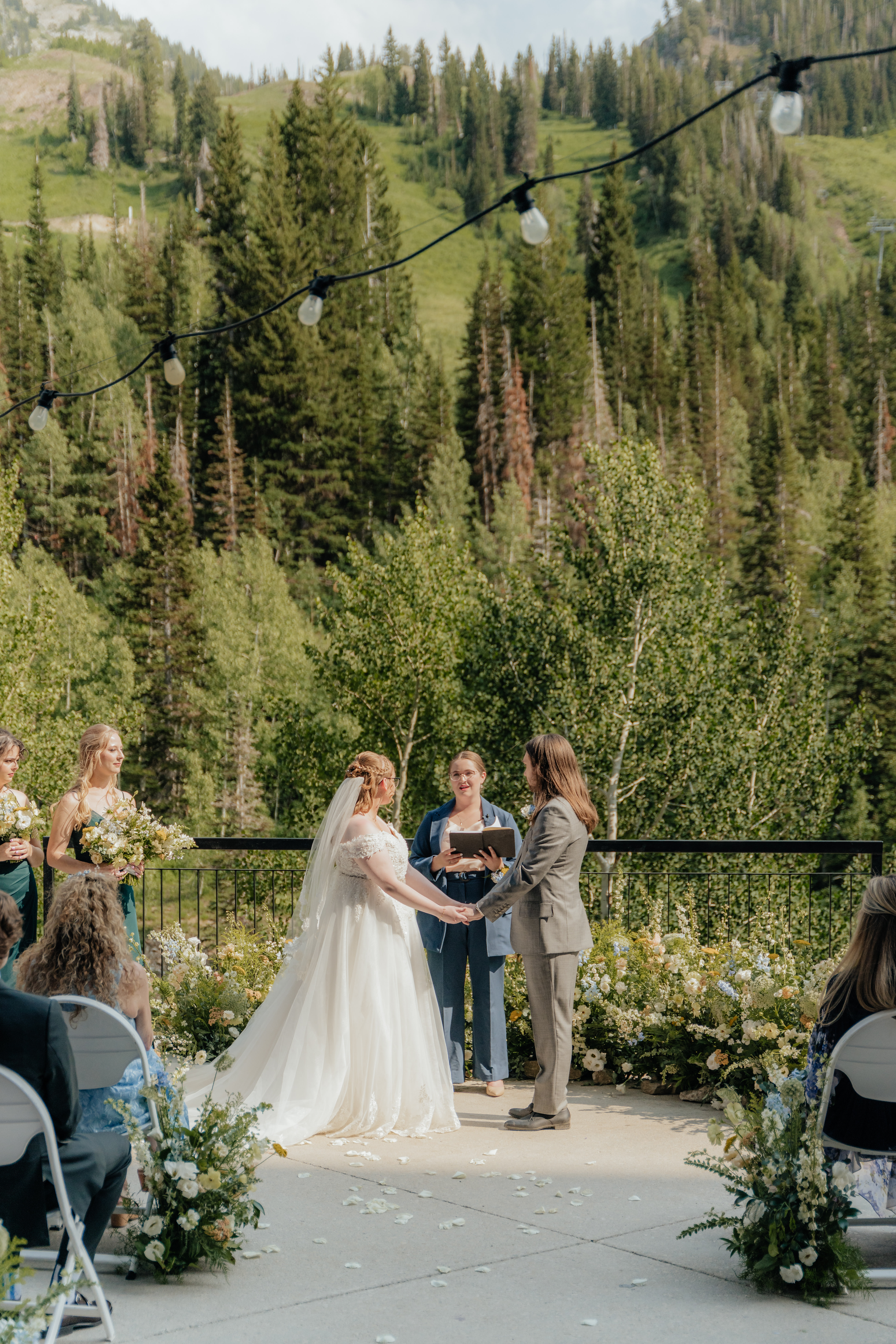 A wedding couple looks lovingly at each other while standing inside a nest of wildflowers and mountain trees as their backdrop. In this photo they are sharing their wedding vows.