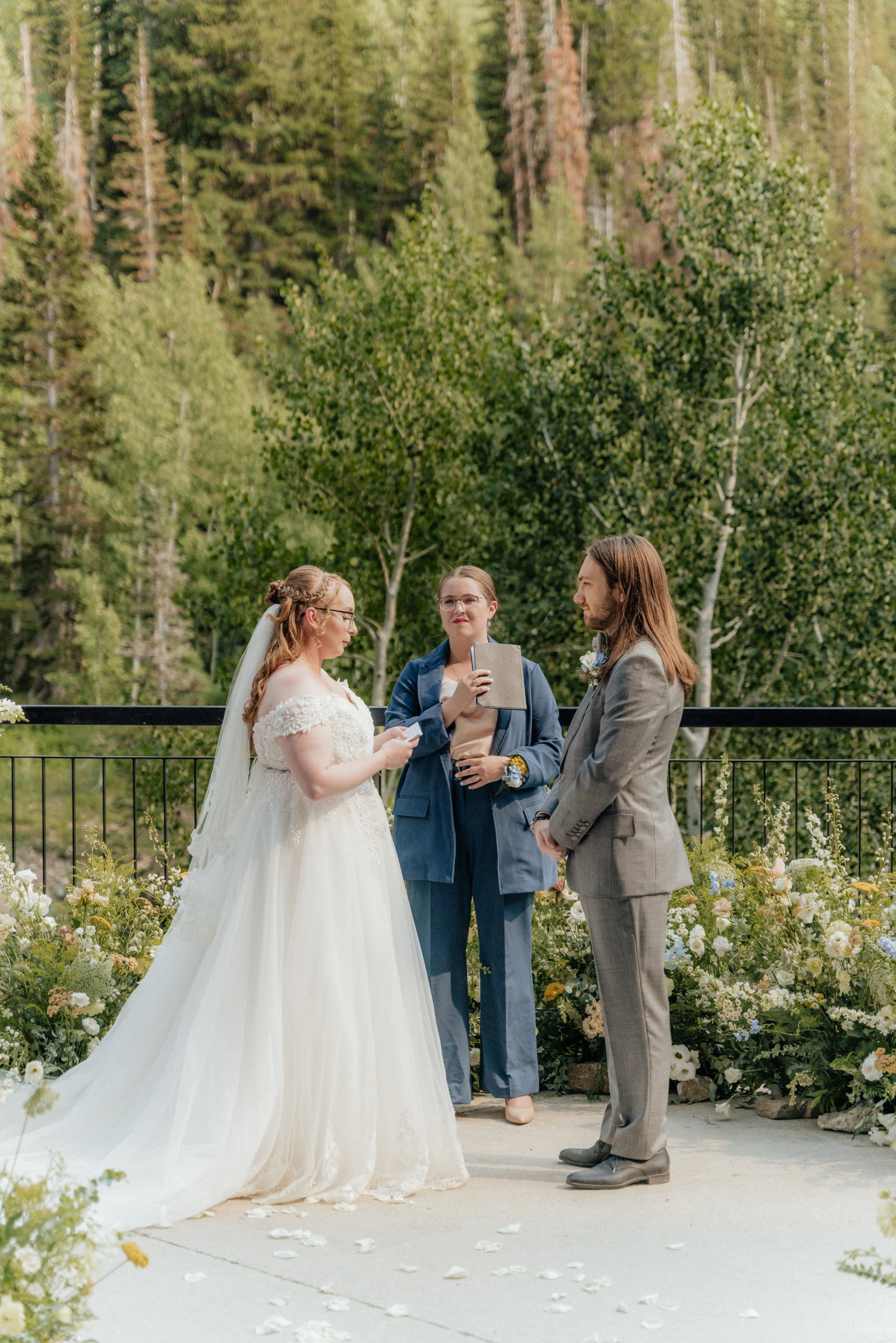A wedding couple looks lovingly at each other while standing inside a nest of wildflowers and mountain trees as their backdrop. In this photo they are sharing their wedding vows.