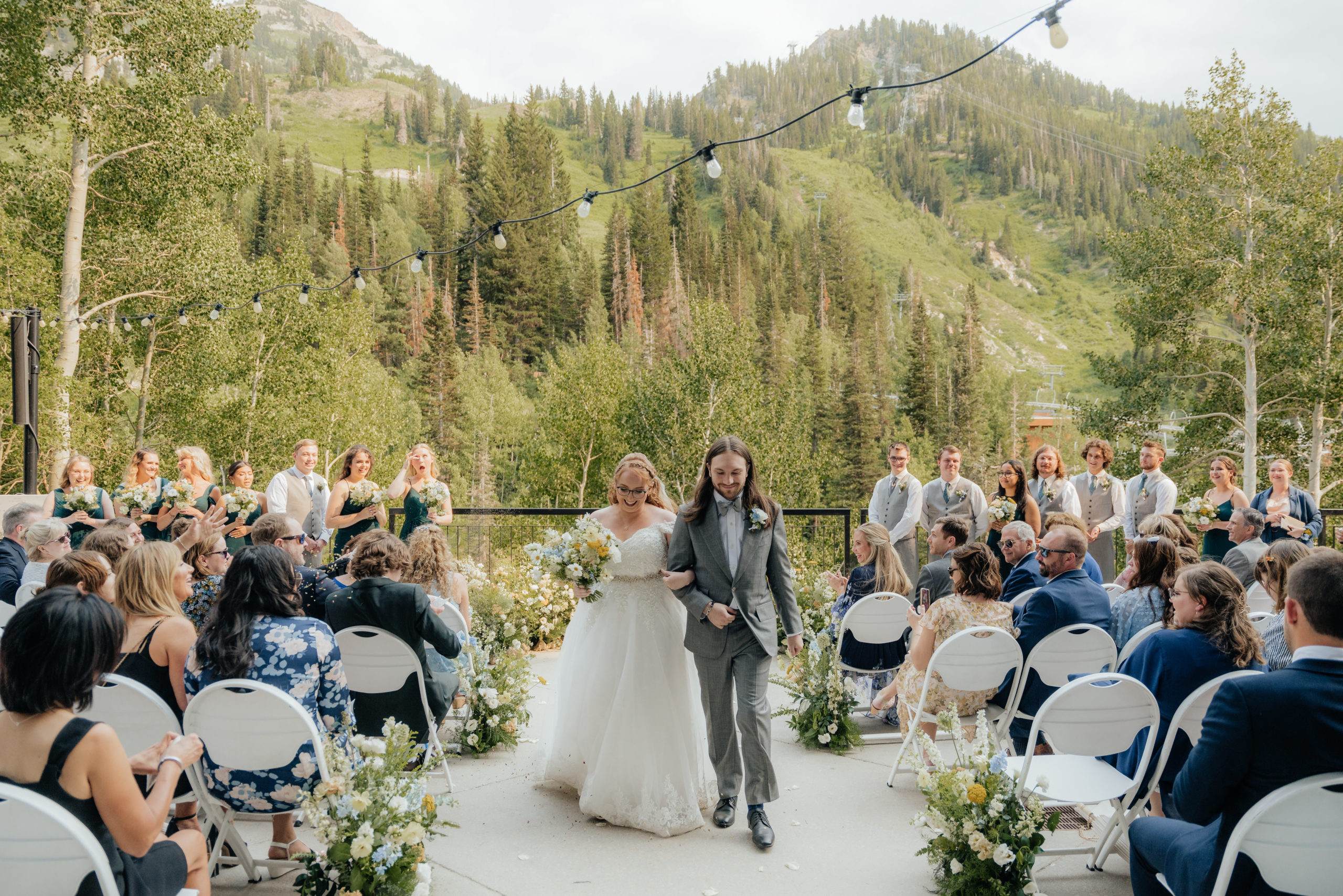 A wedding couple walks excitedly away from a nest of wildflowers and mountain trees as their backdrop after sharing their wedding vows.