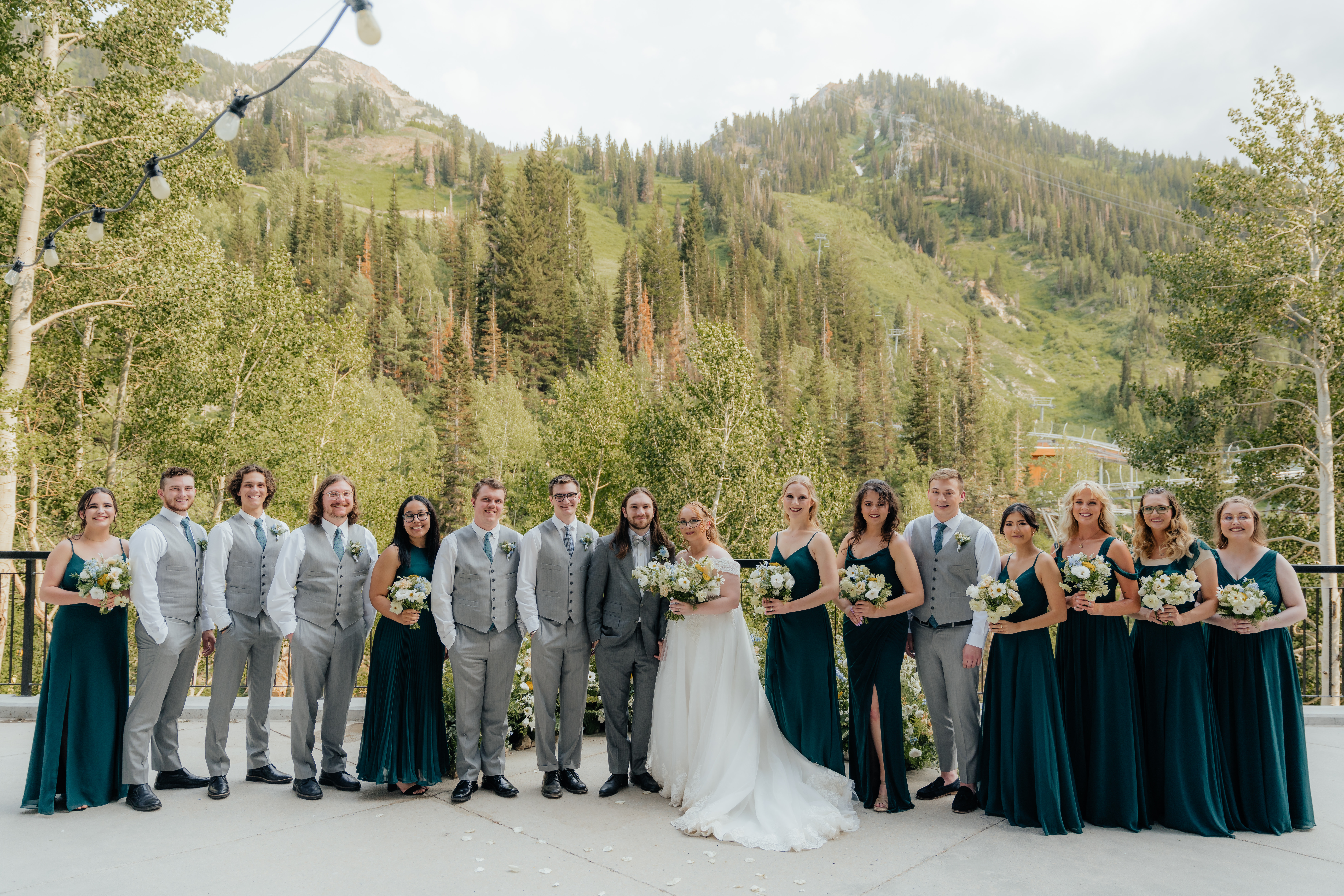 A wedding party of people in deep green dresses and grey suites smile for the camera while standing in front of a nest of wildflowers and mountain trees as their backdrop.