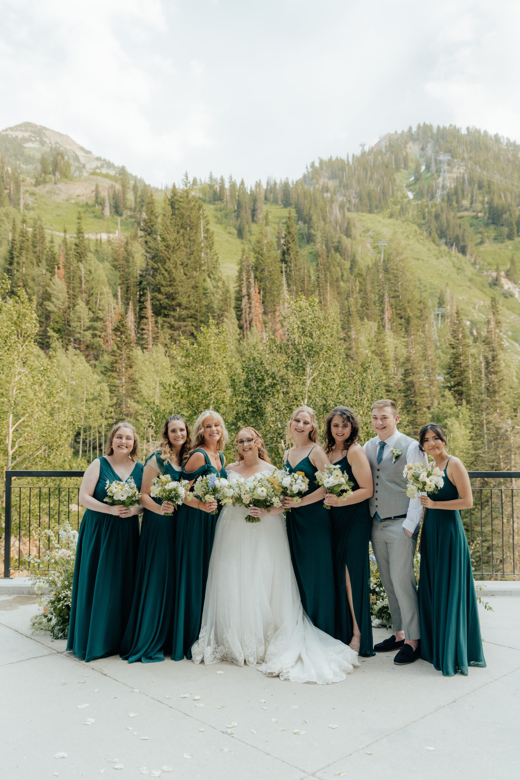 A wedding party of girls in deep green dresses and one guy in a grey suite, smiles for the camera while standing in front of a nest of wildflowers and mountain trees as their backdrop.