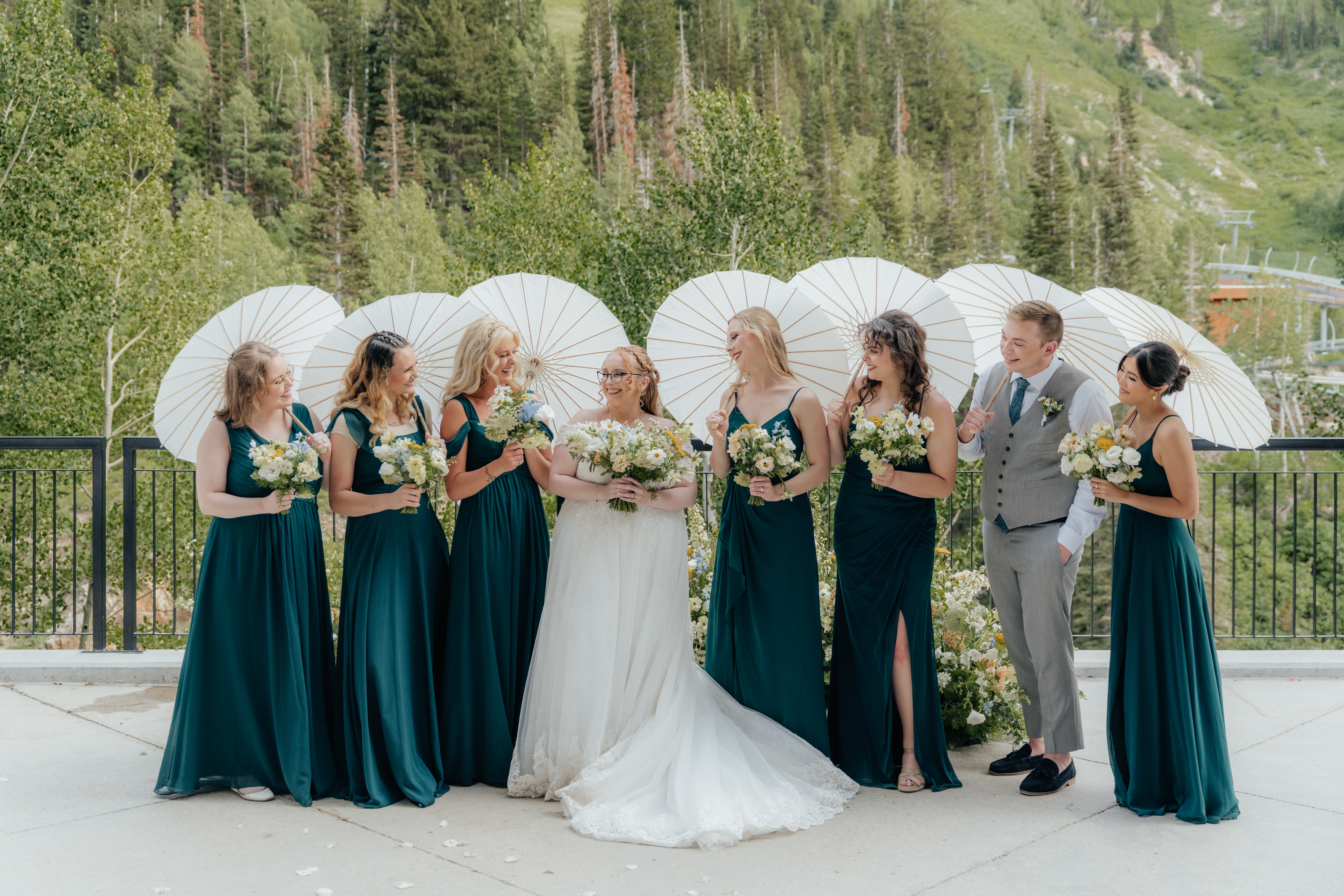 A wedding party of girls in deep green dresses holding white parasols and wildflower wedding bouquets, smile for the camera while standing in front of a nest of wildflowers and mountain trees as their backdrop.