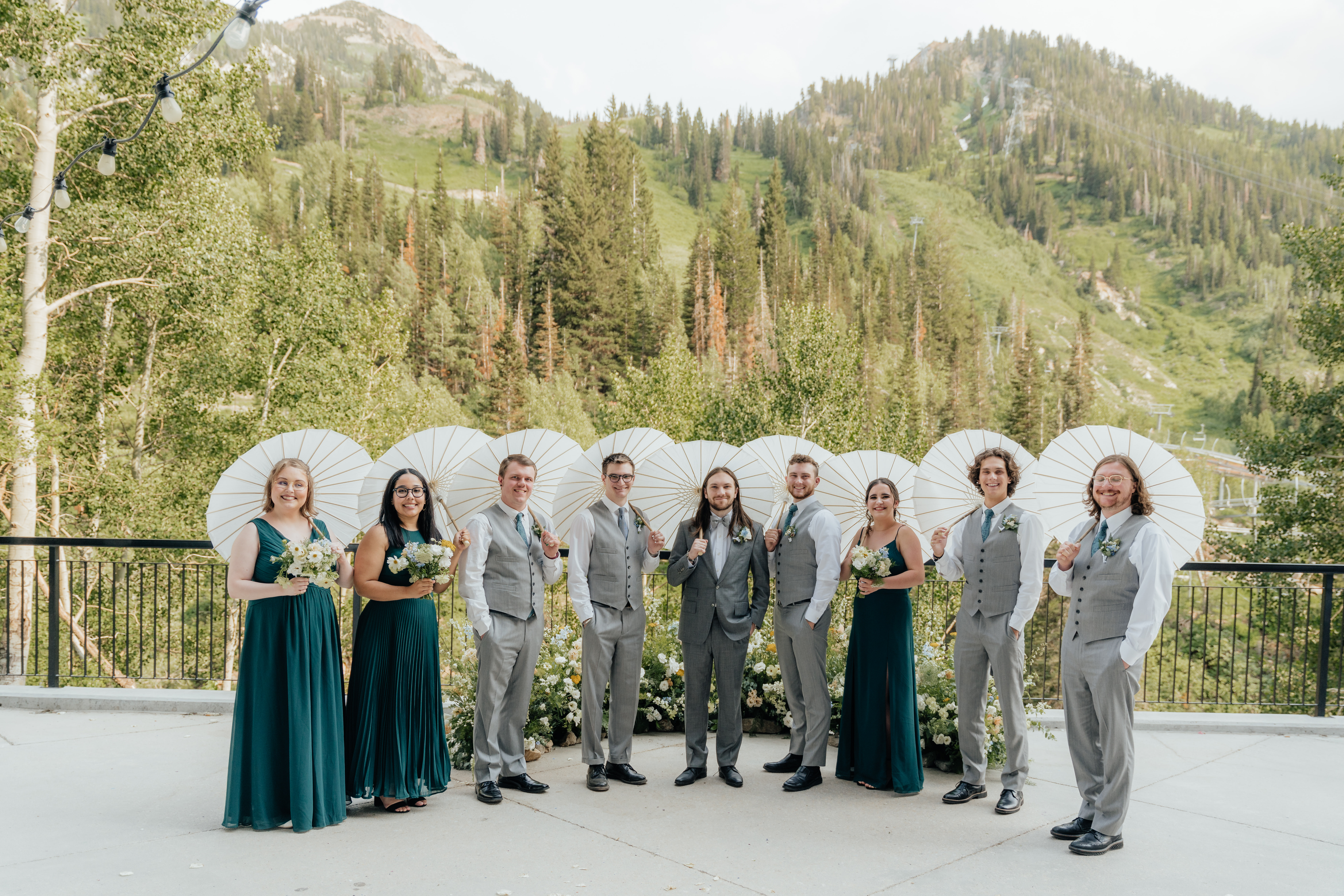 A wedding party of girls in deep green dresses and one guy in a grey suite and holding white parasols, smile for the camera while standing in front of a nest of wildflowers and mountain trees as their backdrop.