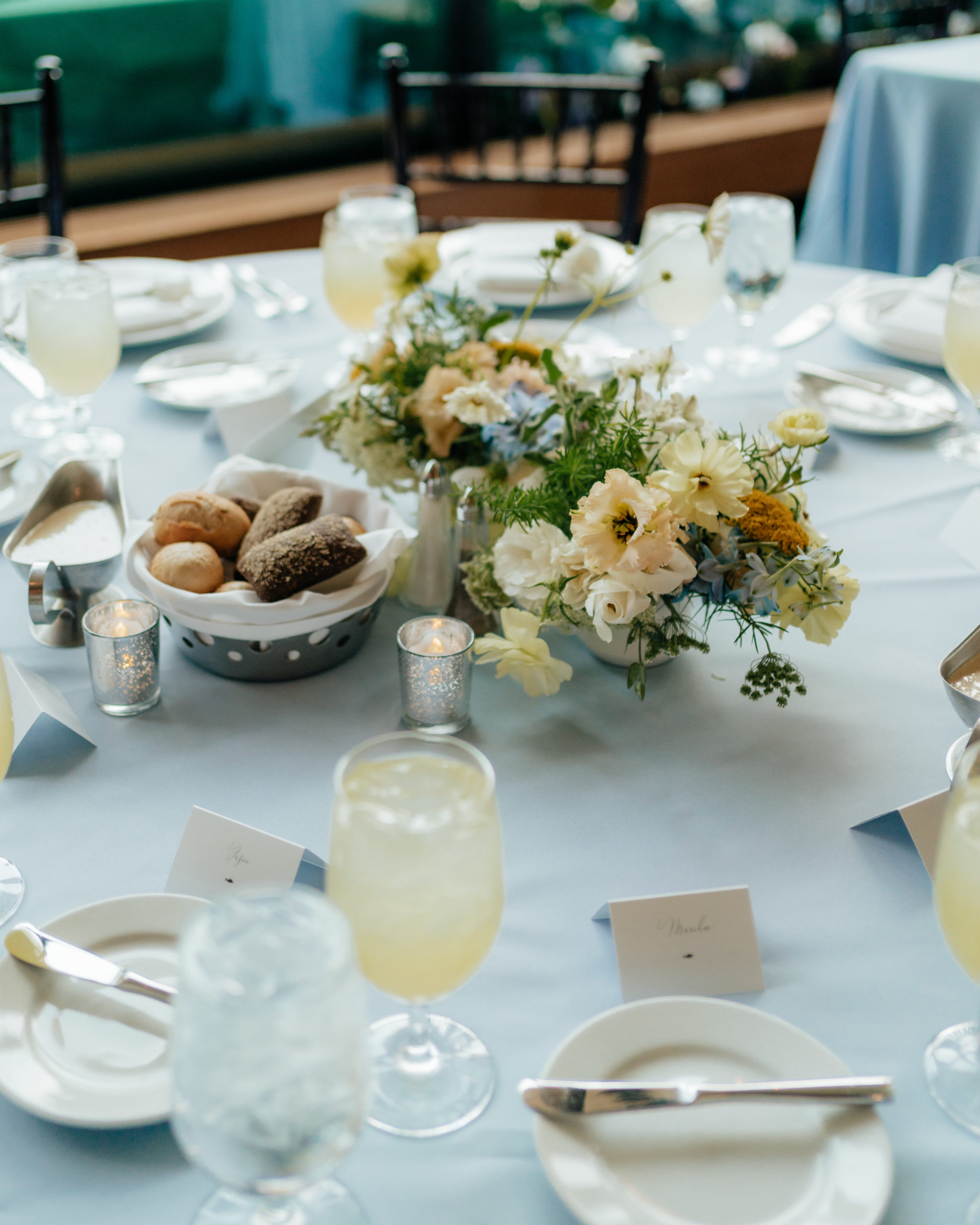 A pretty wedding centerpiece sits on a white table linen. The centerpiece has pops of soft pastel colors like light blue and yellow, giving off a wildflower vibe. Next to the flowers is a basket of rolls and light yellow lemonade in a glass.