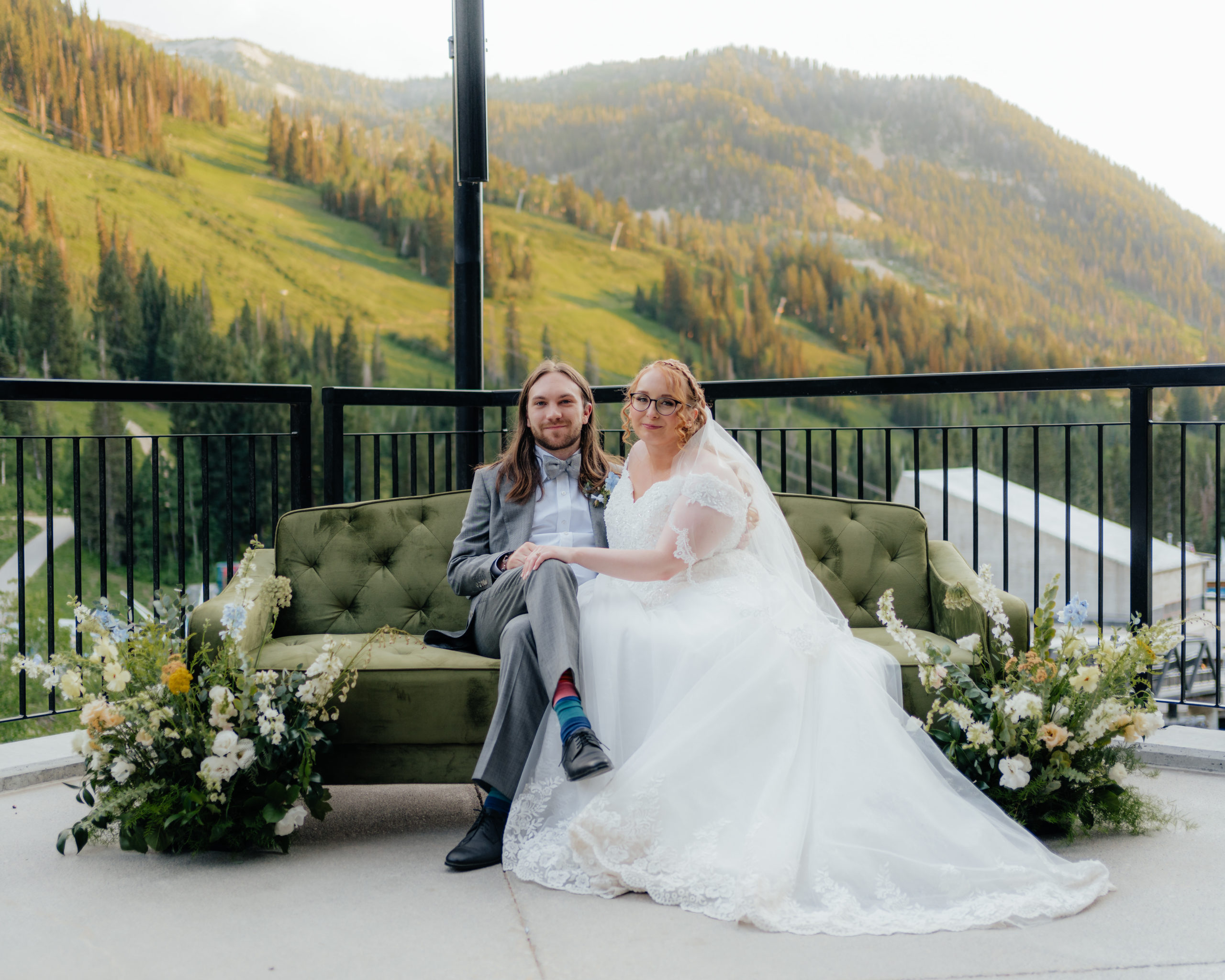 A wedding couple looks lovingly at the camera while sitting on a pretty green couch, wildflowers to their side and mountain trees as their backdrop.