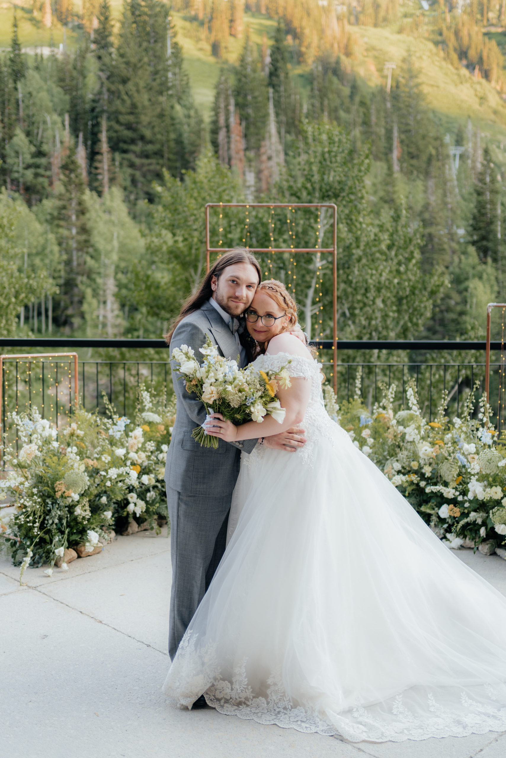 A wedding couple looks lovingly at the camera while standing inside a nest of wildflowers and mountain trees as their backdrop.