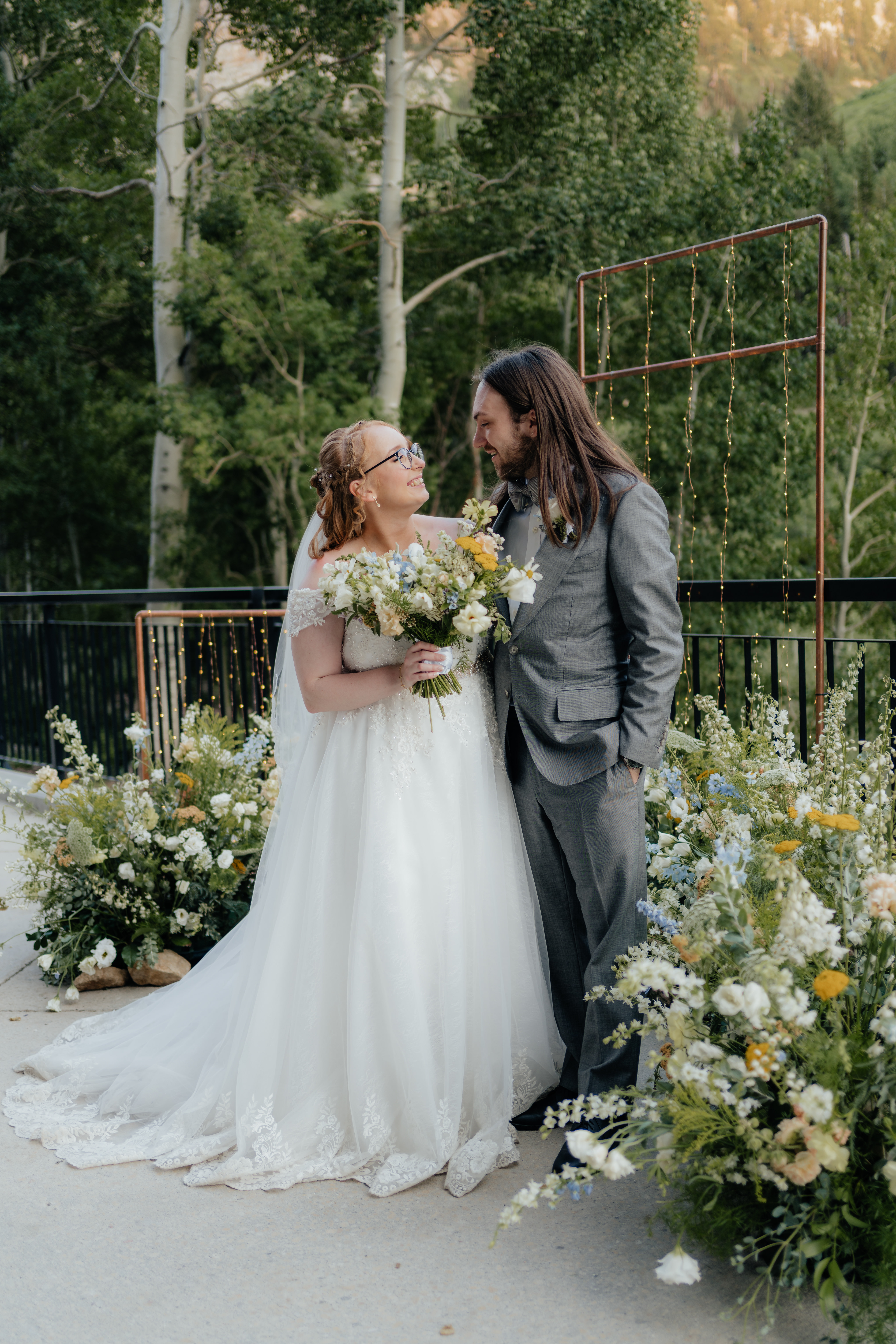 A wedding couple looks lovingly at each other while standing inside a nest of wildflowers and mountain trees as their backdrop.